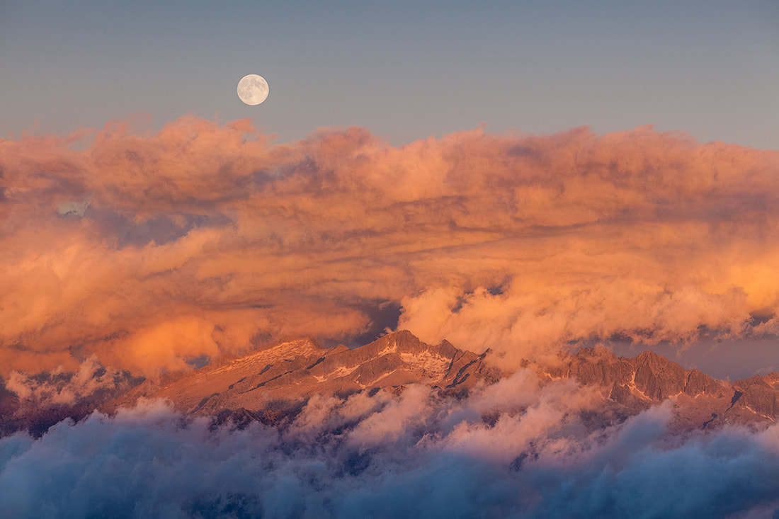 La luna piena spunta al tramonto dalla catena dell'Adamello, circondato da nuvole